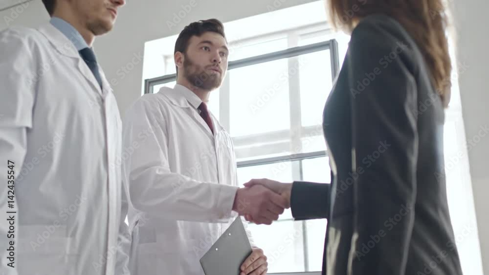 Low angle panning shot of businesswoman in suit shaking hands of scientists in lab coats during meeting, then listening to them reading report on latest experiment 