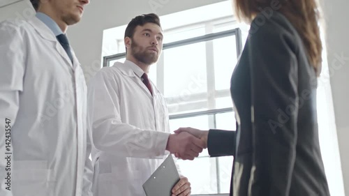 Low angle panning shot of businesswoman in suit shaking hands of scientists in lab coats during meeting, then listening to them reading report on latest experiment  photo
