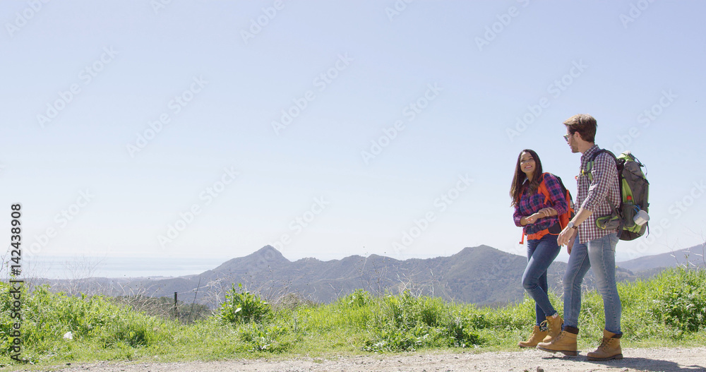 Young laughing couple wearing backpacks and walking down road while hiking in mountains in summer time. 