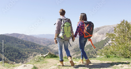 Back view of young couple with backpack holding hands and enjoying view of mountains in summer time. 