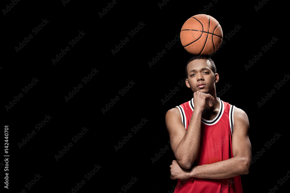 Young confident basketball player posing with ball on head on black