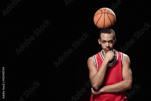 Young smiling basketball player standing with ball on head on black