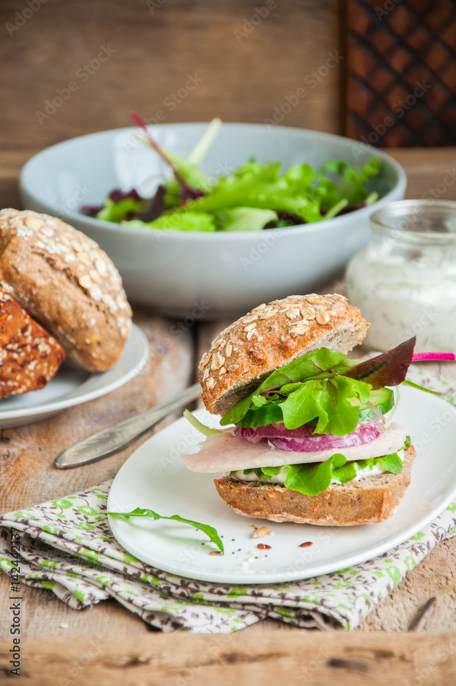 Sandwich with salad herbs chicken ham red onion seeds and spicy sour cream sauce served on a white plate. Wooden background.