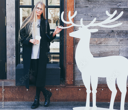 Blond girl hipster in round glasses with black outfit resting and drinking coffee from a paper cup at the entrance to a fashionable cafe posing with a figure of a white deer photo