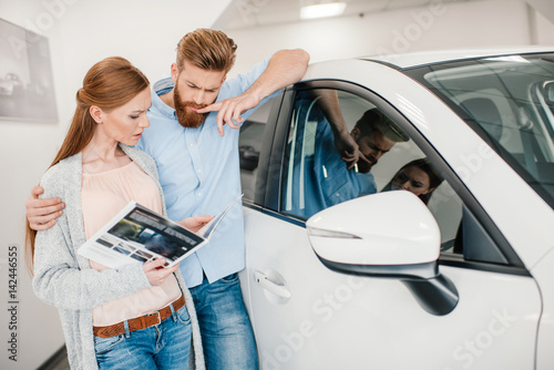 Valokuva pensive couple holding catalog and choosing car in dealership salon