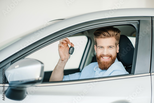 Smiling bearded young man sitting in new car and holding key