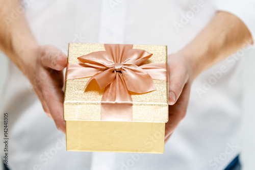 Male hands holding a gift box. Present wrapped with ribbon and bow. Christmas or birthday golden paper package. Man in white shirt.