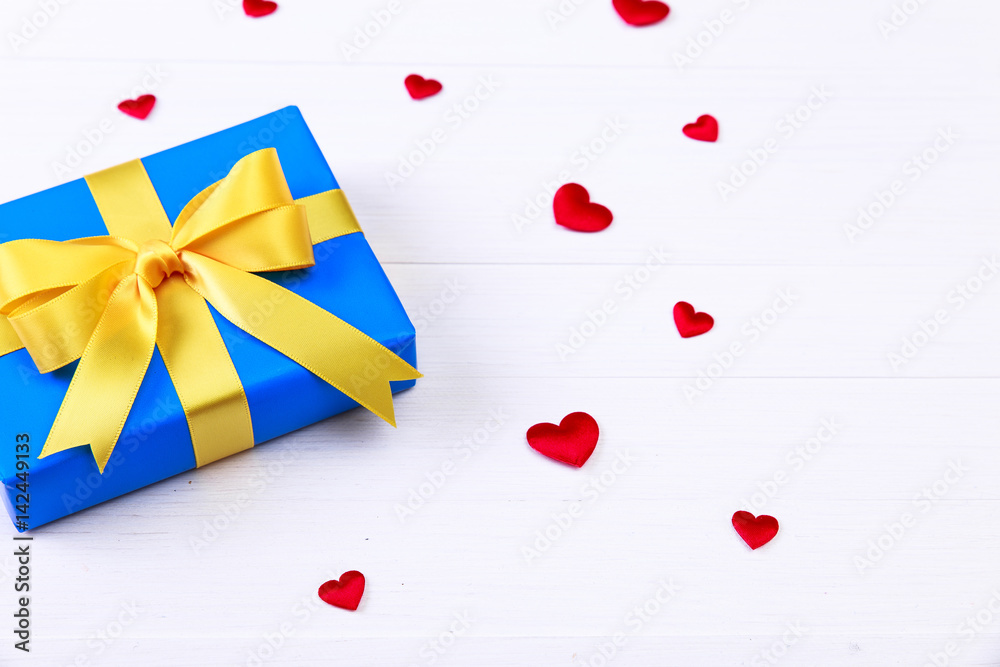 Gift box with red satin hearts. Present wrapped with yellow ribbon. Christmas or birthday blue package. On white wooden table. Gift box with red satin hearts. Present wrapped with yellow ribbon. Christmas or birthday blue package. On white wooden table.