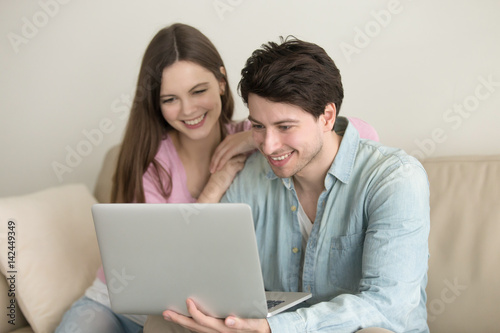 Young happy couple sitting with laptop computer, making video call, hand on shoulder, online shopping, looking at screen, social networking, events guide, browsing, planning vacation, talking, working © fizkes