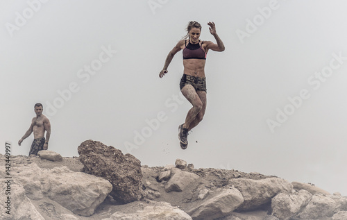 Strong beautiful muscular female athlete covered in mud jumping of a rough rocky ledge capture in mid air. 