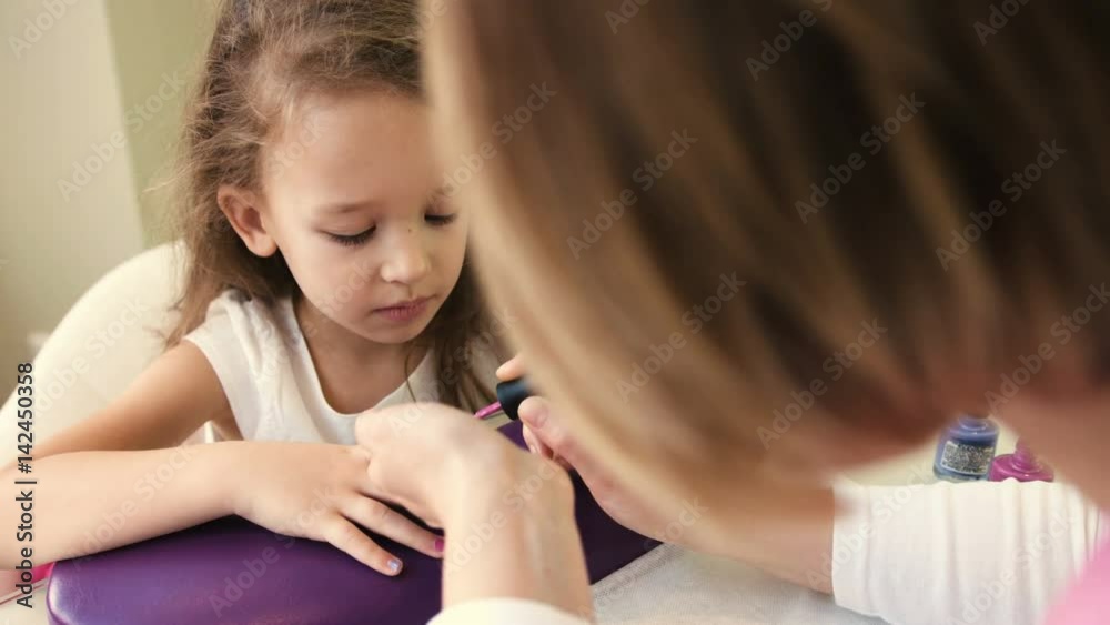 Nail master doing manicure for pretty little girl in professional salon. Close up view of manicure artist applying gel polish Nail master doing manicure for pretty little girl in professional salon. Close up view of manicure artist applying gel polish