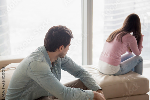 Young couple sitting apart indoors after quarrel. Offended girl ignoring boyfriend, looking away, serious man thinking about problem in relationships, family conflict, misunderstanding between teens