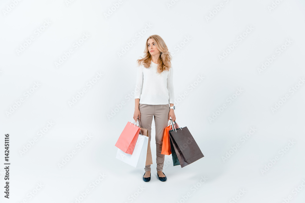 Tired blonde woman holding shopping bags and looking away on grey