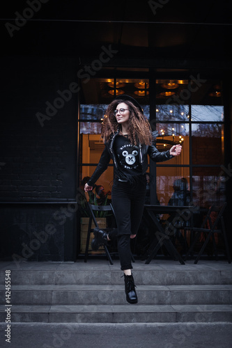 Portrait of a beautiful girl with brown eyes in glasses in a black hat and coat with fur jumping from the stairs of a city cafe and looking away. The girl is like Harry Potter