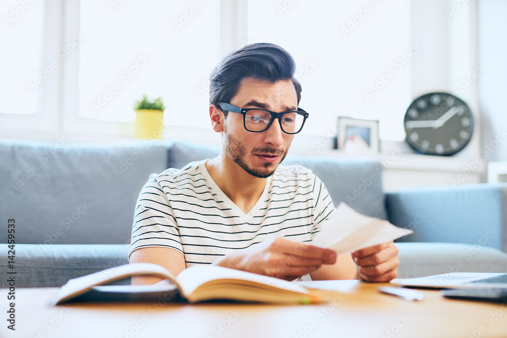 Young man paying bills at home looking surprised Young man paying bills at home looking surprised
