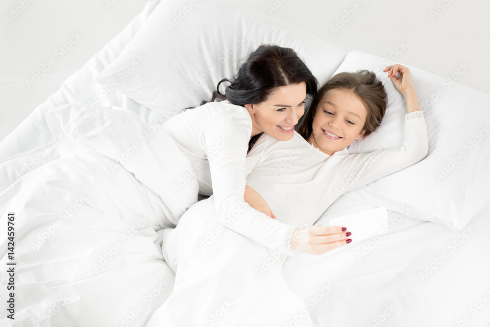 Adorable daughter and mother taking selfie on smartphone while lying on bed Adorable daughter and mother taking selfie on smartphone while lying on bed