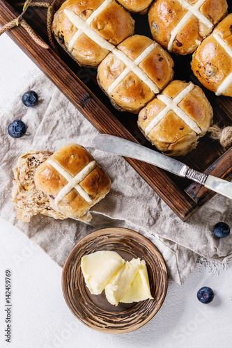 Hot cross buns in wooden tray served with butter, fresh blueberries, knife and jug of cream on textile napkin over white texture concrete background. Top view, space. Easter baking.
