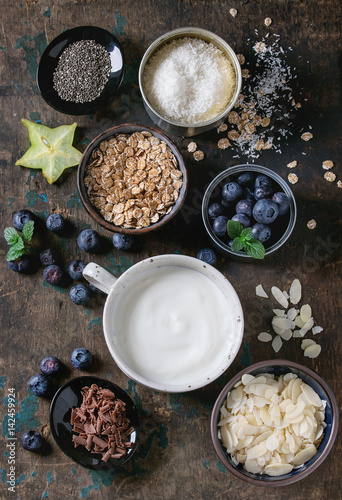 Ingredients for making smoothie for healthy breakfast. Bowls of yogurt, blueberries, granola, almond chia seeds, coconut, milk, chocolate, mint, carambola over dark wooden background. Top view, space