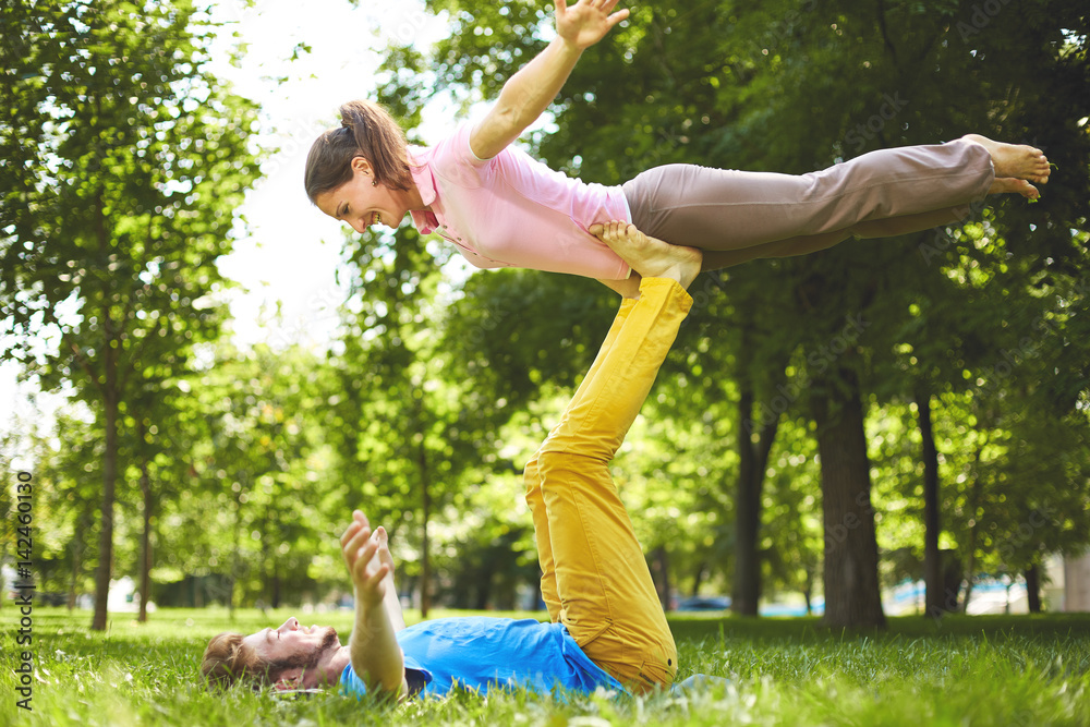 Fit young couple doing acro yoga in park. Man lying on grass and balancing  woman in
