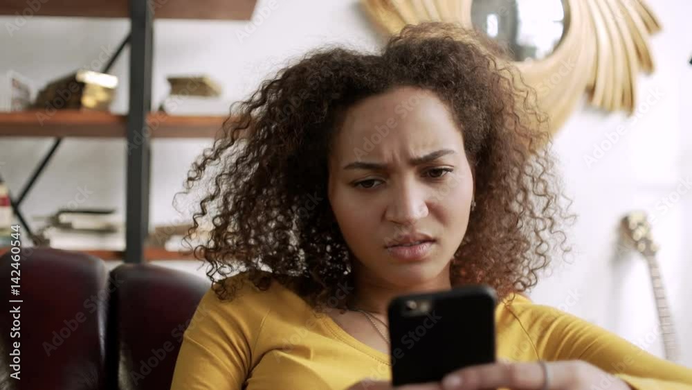 young curly latin woman frown using mobile phone because get bad message and fake new in internet or social network typing messages sitting at home on comfort sofa at home during sunny day