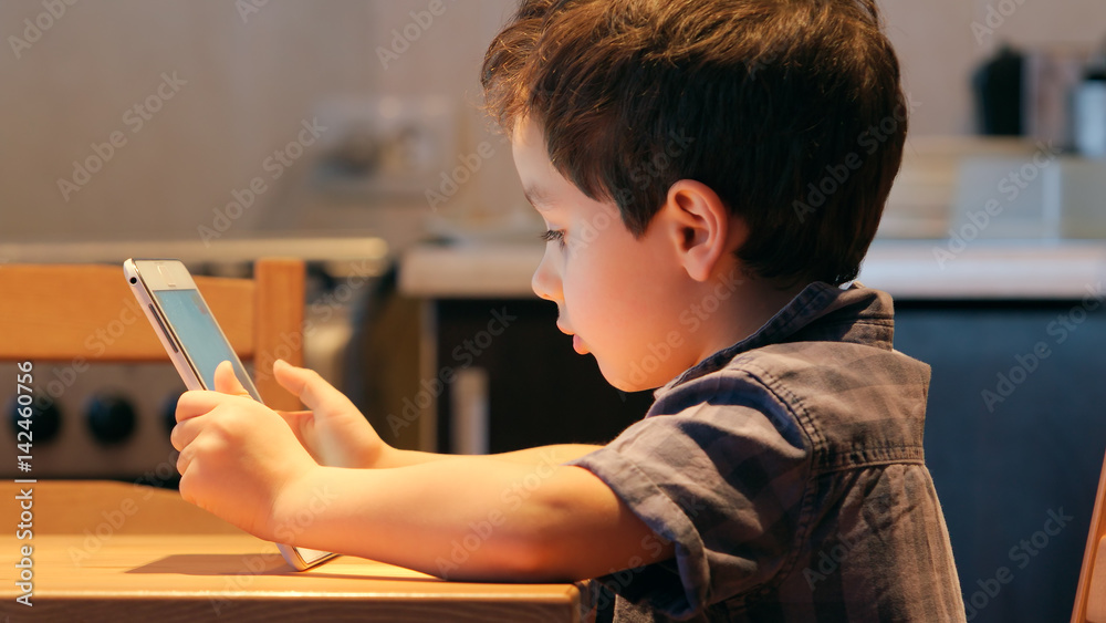 Cute little child uses a tablet PC at a table at home. Casual clothes. Side view Cute little child uses a tablet PC at a table at home. Casual clothes. Side view