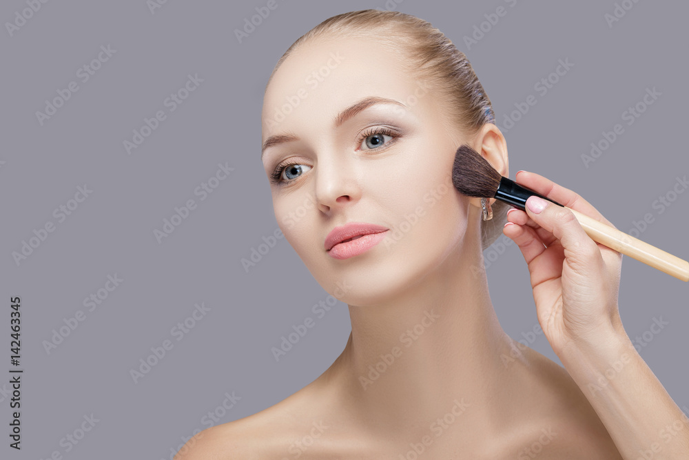 beautiful blond woman holding makeup brush on a gray background. woman applying blusher