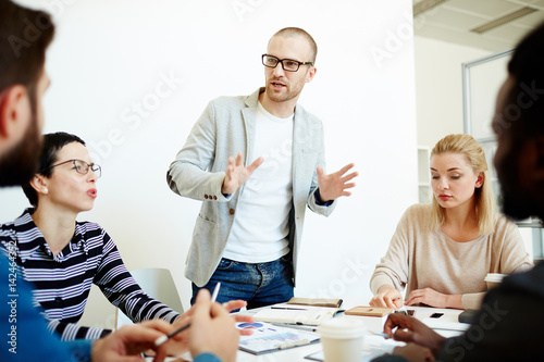 Confident team leader in eyeglasses leading informal meeting in boardroom while his colleagues listening to him with attention, notebooks, pens and documents lying on table