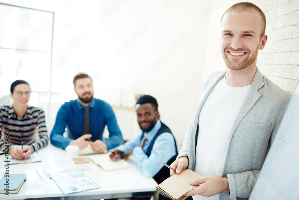 Portrait of cheerful young businessman in casualwear with pen and notebook in hands looking at camera while leading business meeting for his subordinates Portrait of cheerful young businessman in casualwear with pen and notebook in hands looking at camera while leading business meeting for his subordinates