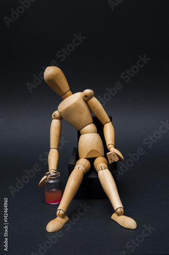 Person represented by a wooden dummy looking rather dizzy or drunk while sitting on an armchair with an opened bottle of alcoholic drink aside. 