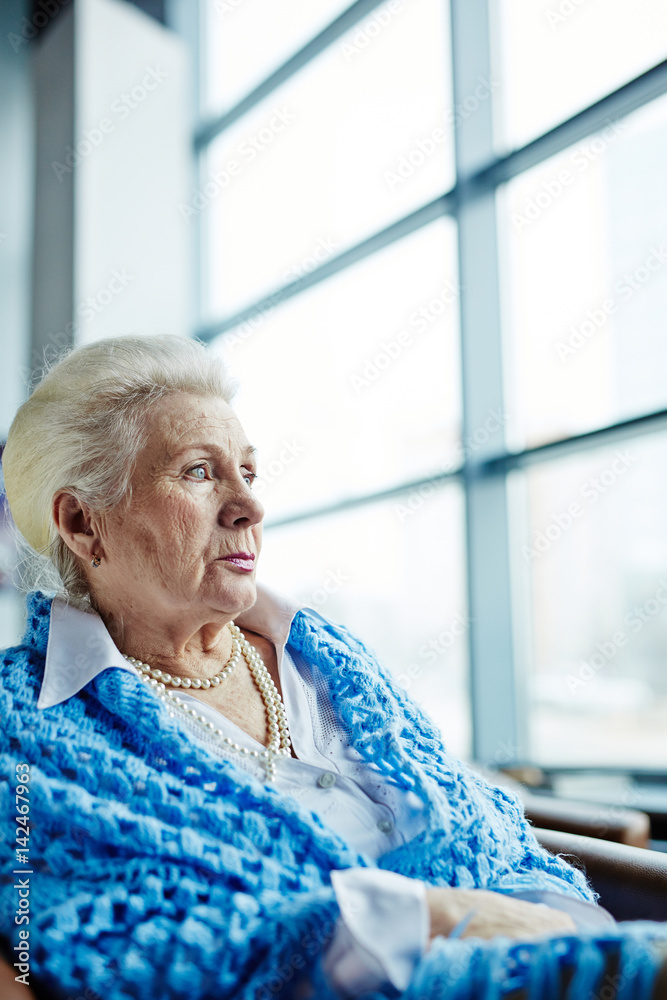 Waist-up portrait of beautiful elderly woman with pearl necklace and cornflower blue knitted shawl sitting on armchair and looking out panoramic window Waist-up portrait of beautiful elderly woman with pearl necklace and cornflower blue knitted shawl sitting on armchair and looking out panoramic window