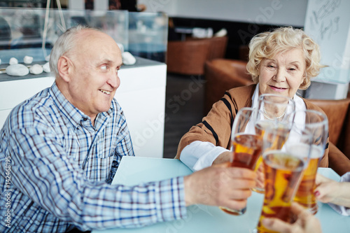 Profile view of handsome senior man looking with toothy smile at his pretty female friend while clanging beer glasses together