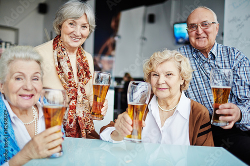 Waist-up portrait of four elderly people gathered together in pub and posing for photography with beer glasses in hands