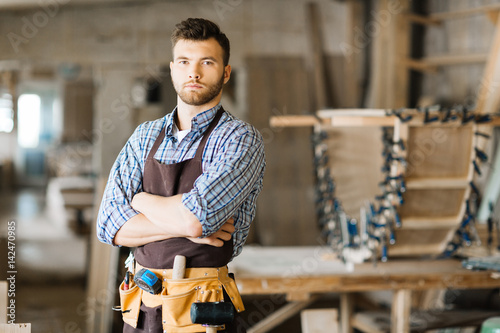 Portrait of serious young woodworker wearing leather tool belt posing for photography while standing in workshop with arms crossed photo