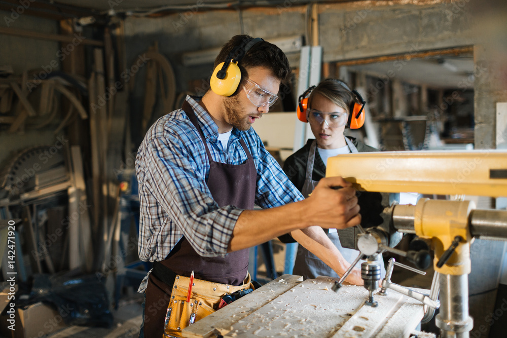 Skillful woodworker in eye and ear protectors explaining his female apprentice how to use drill press, she listening to him with attention Skillful woodworker in eye and ear protectors explaining his female apprentice how to use drill press, she listening to him with attention