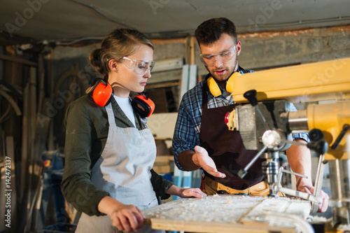 Waist-up portrait of attractive apprentice standing at drill press and listening to her male mentor with attention, he explaining her operating principle of machine