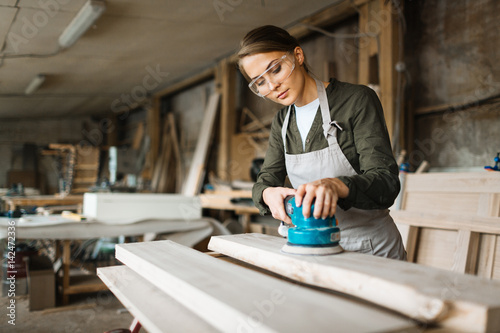 Assiduous young woodworker wearing safety glasses and apron while using electric sander in spacious workshop, waist-up portrait photo