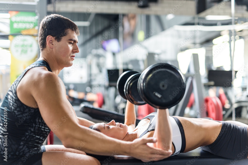 Portrait of muscular personal instructor helping beautiful woman doing weightlifting exercise with big dumbbells on bench in modern gym Portrait of muscular personal instructor helping beautiful woman doing weightlifting exercise with big dumbbells on bench in modern gym