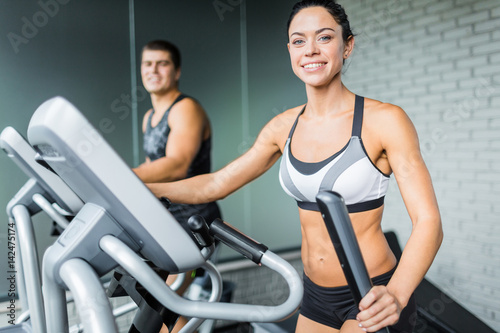 Portrait of beautiful  sportive brunette woman exercising using elliptical machine  next to fit man, both smiling to camera during workout in modern gym © pressmaster