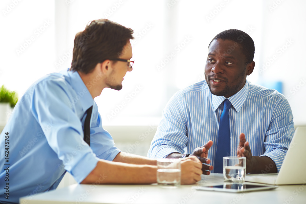 Waist-up portrait of Afro-American businessman in formalwear discussing promising project with his male colleague while sitting in meeting room Waist-up portrait of Afro-American businessman in formalwear discussing promising project with his male colleague while sitting in meeting room