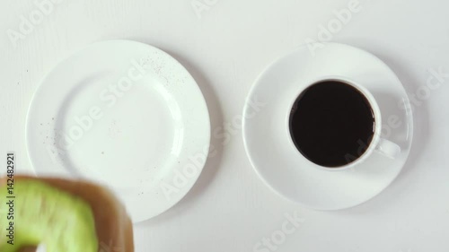 A female hand takes a donut covered with green icing with cinnamon from a saucer and a cup of coffee, sips down coffee and bites a donut. Top view