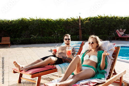 Friends smiling, drinking cocktails, lying on chaises near swimming pool. © Cookie Studio