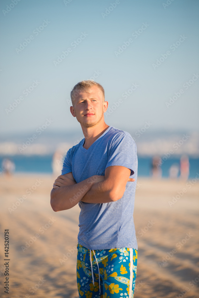 Guy with white hair dressed in shorts stands on the beach near the sea - obrazy, fototapety, plakaty 