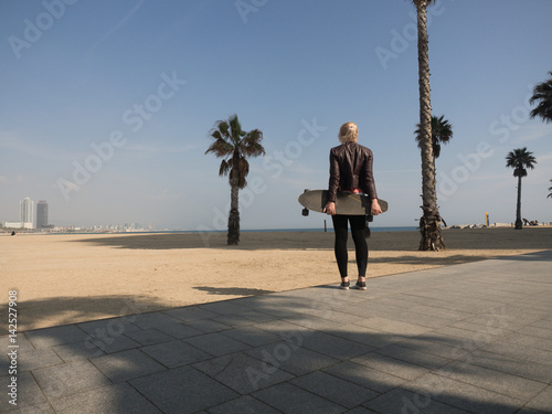 Photography Young modern woman with leather jacket holding a skateboard at the beach in fron