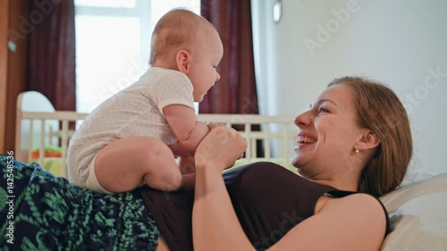 Young Mother is helping her cute baby to sit up at home. Baby girl is sitting on mother s stomach and smiling photo