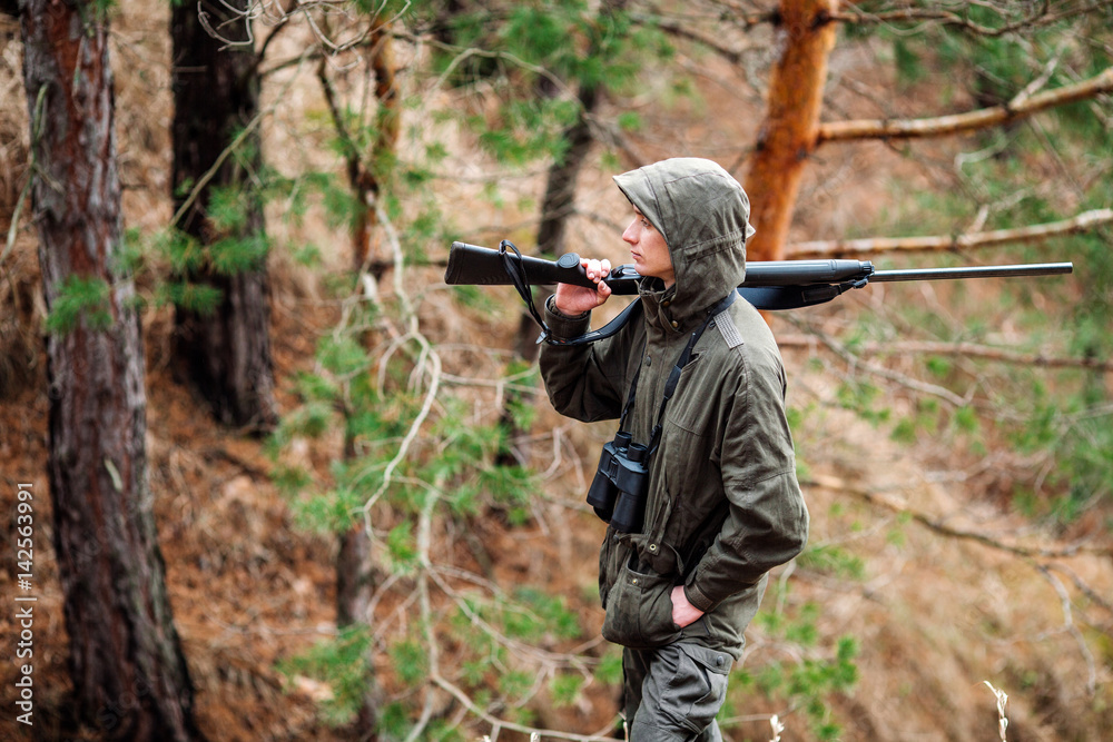 Naklejka premium male hunter with binoculars ready to hunt, holding gun and walking in forest.