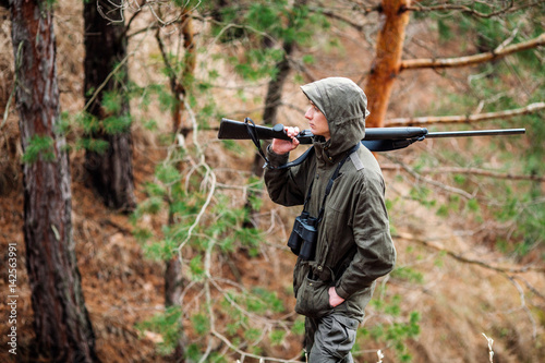 male hunter with binoculars ready to hunt, holding gun and walking in forest.