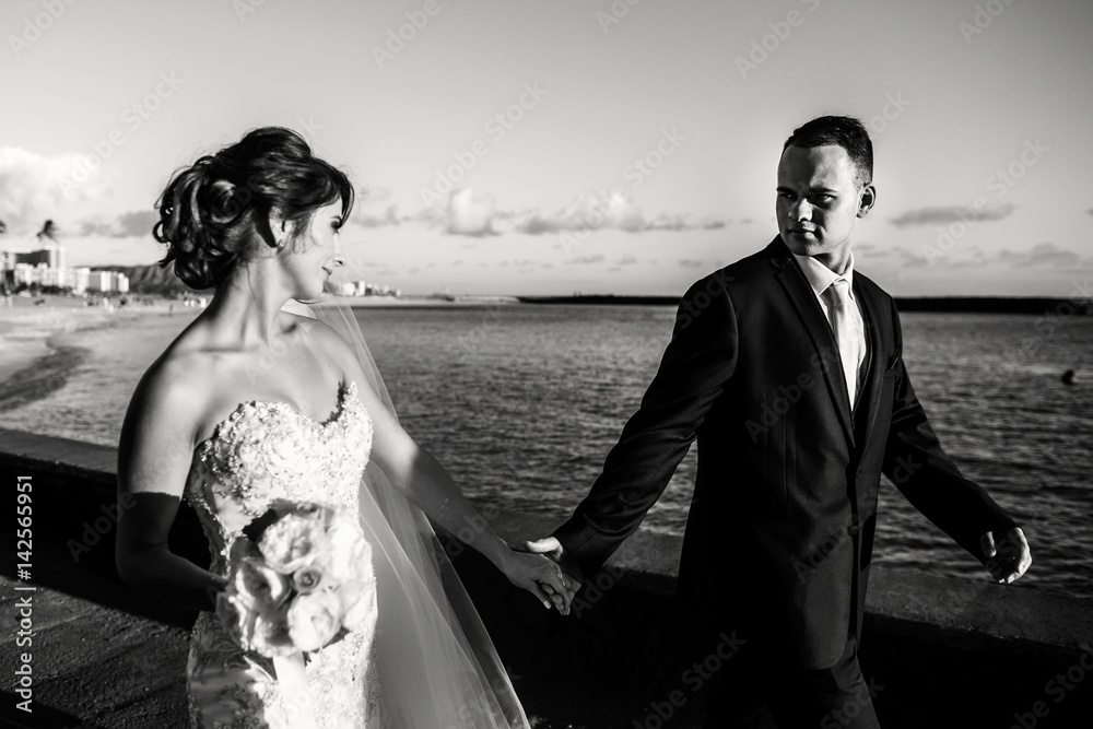 Groom looks over his shoulder at bride walking along ocean shore