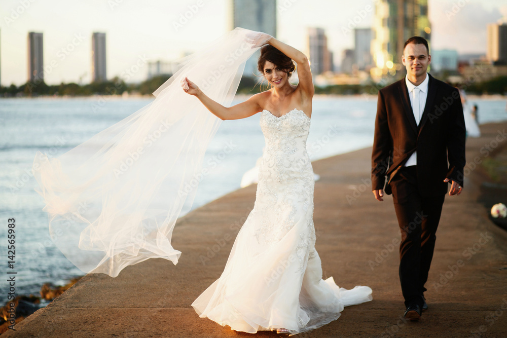 Bride fixes her veil walking with groom along ocean shore