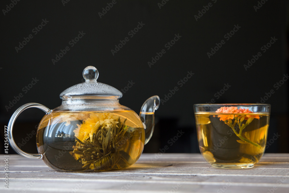 a glass tea pot with Flower Chinese tea and a cap of green tea on wooden table in front of dark background a glass tea pot with Flower Chinese tea and a cap of green tea on wooden table in front of dark background