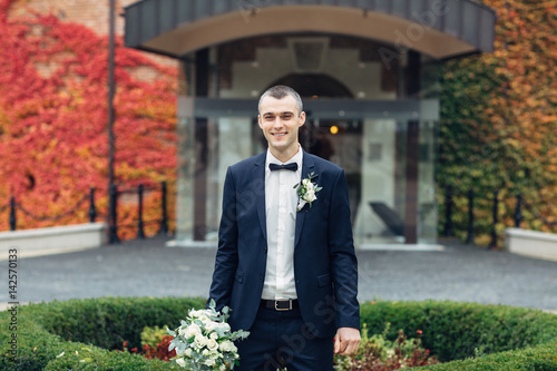Groom in dark blue suit stands with wedding bouquet before entrance to the hotel